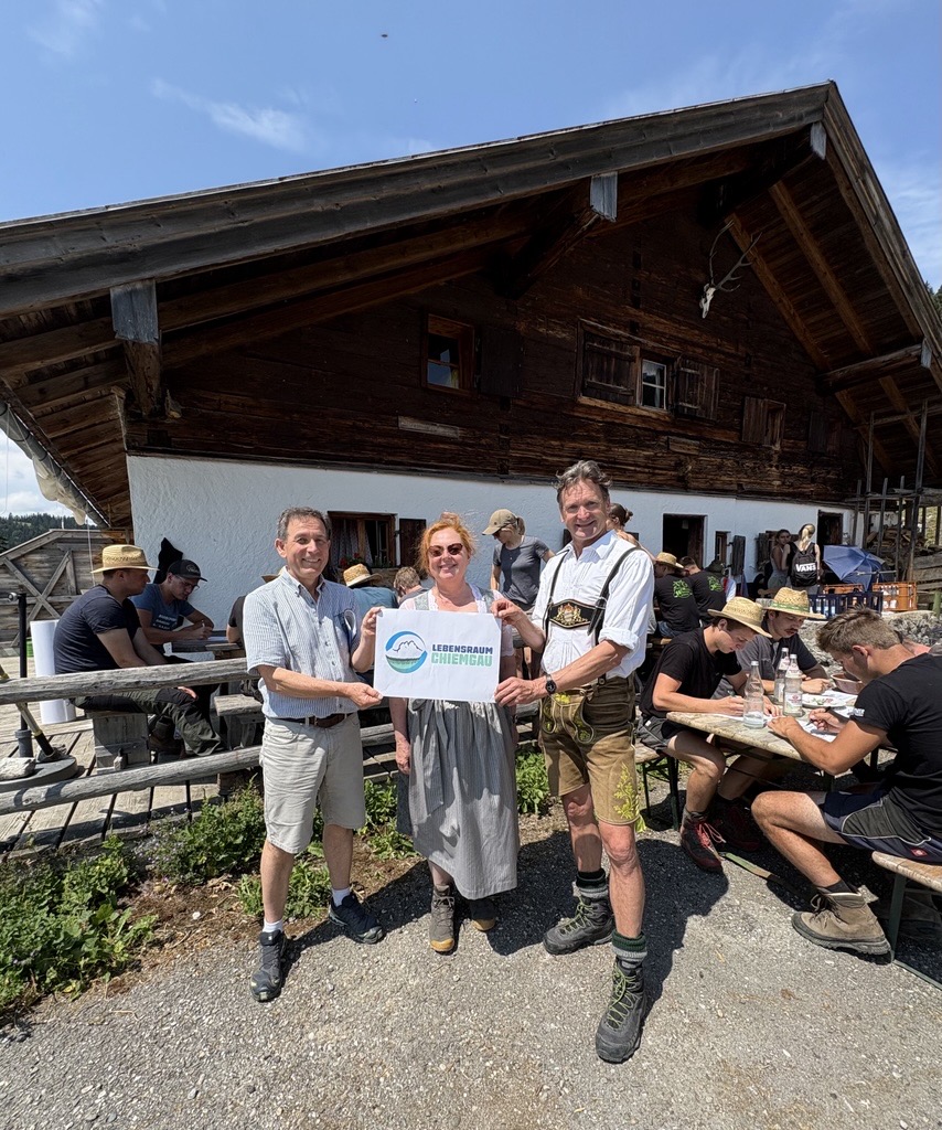 Die beiden Projektleiter stehen mit einer weiteren Projektbeteiligten vor einer Almhütte und halten das Logo von Lebensraum Chiemgau